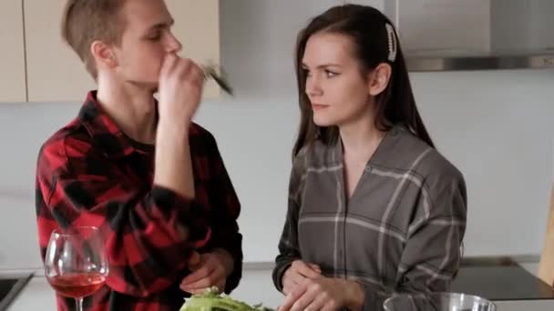 Jovem casal bonito em camisas xadrez estão cozinhando em casa na cozinha. Uma mulher e um homem cortam legumes e fazem uma salada de pimenta, tomate, rabanete em um prato de vidro transparente. . — Vídeo de Stock