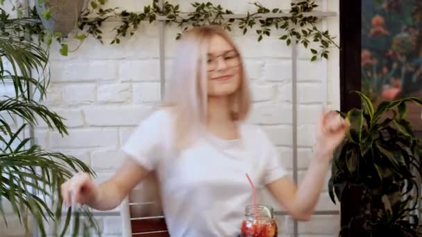 Νεαρή όμορφη γυναίκα ξανθιά σε γυαλιά που χορεύουν στη μουσική από ένα smartphone σε ένα τραπέζι καφέ. — Αρχείο Βίντεο