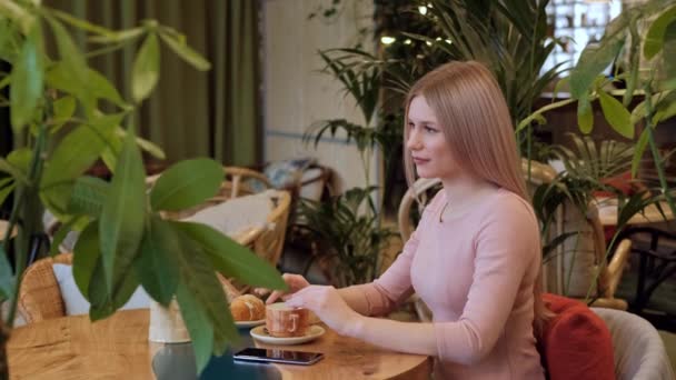 Jovem mulher loira bonita em um vestido rosa sentado a uma mesa em um café e beber chá . — Vídeo de Stock