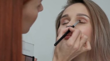 Makyaj sanatçısı hafif bir monofonik arka plan üzerinde makyaj güzel sarışın kız yapar. Gözleri maskara ile boyar, fırça ile toz uygular.