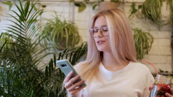 Jonge mooie vrouw blonde in glazen dansen op de muziek van een smartphone in een café tafel. — Stockvideo