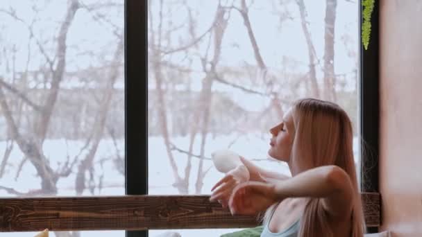 Junge schöne Blondine in Sportbekleidung, die am Fenster sitzt. streckt sich und trinkt Limonade aus einem Glas. — Stockvideo