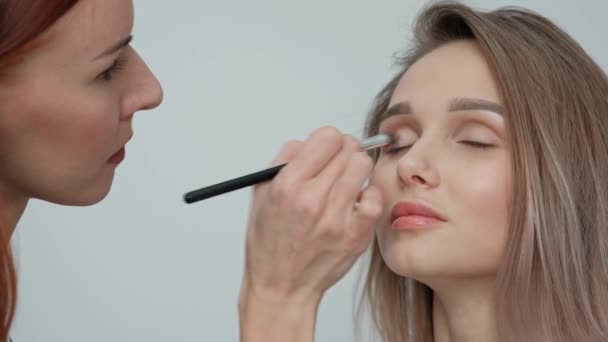 Makeup artist gör makeup vacker blond flicka på en ljus monofoniska bakgrund. Målar ögon med mascara, applicerar puder med en borste. — Stockvideo