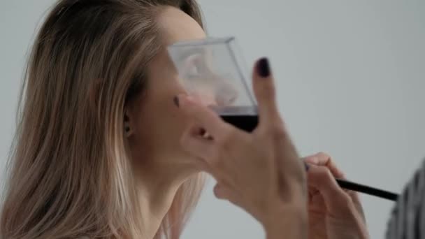 Make-up artiest maakt make-up mooi blond meisje op een lichte monofische achtergrond. Schildert de ogen met mascara, past poeder toe met een borstel. — Stockvideo