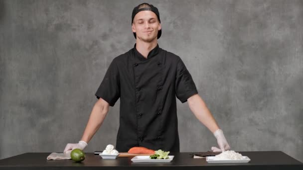 Jonge man chef kok in zwarte kleren op een grijze achtergrond. Keuken tafel, ingrediënten voor Japanse sushi en broodjes. — Stockvideo