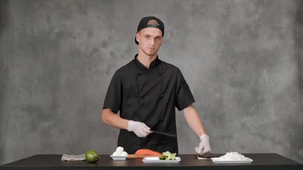 Jeune homme chef cuisinier en vêtements noirs sur un fond gris. Table de cuisine, ingrédients pour sushi japonais et petits pains . — Video