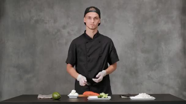 회색 배경에 검은 옷을 입고 요리하는 청년 요리사. 주방 테이블, 일본 초밥과 롤 재료. — 비디오