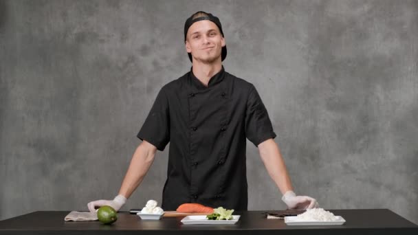 회색 배경에 검은 옷을 입고 요리하는 청년 요리사. 주방 테이블, 일본 초밥과 롤 재료. — 비디오