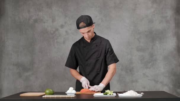 Молодой повар готовит в черной одежде на сером фоне. Кухонный стол, ингредиенты для японских суши и рулонов . — стоковое видео