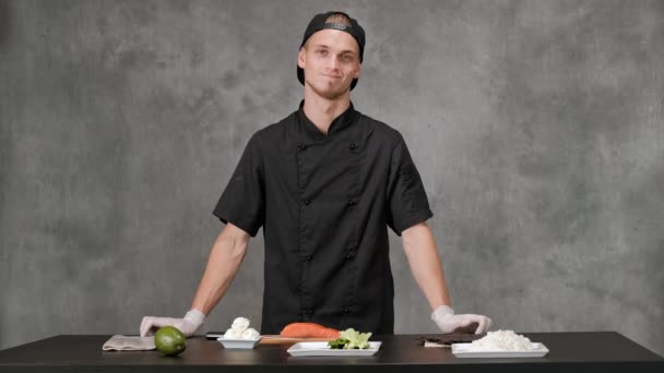 Genç adam şef gri bir arka plan üzerinde siyah giysiler içinde yemek. Mutfak masası, Japon suşi ve rulo için malzemeler. — Stok video
