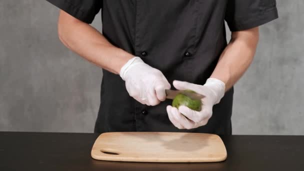 Ung man i svarta kläder på en grå bakgrund peeling, avokado med en kniv. — Stockvideo