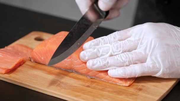 Un jeune chef sushi coupe un filet de saumon fumé avec un couteau sur une table. Mains dans les gants gros plan . — Video