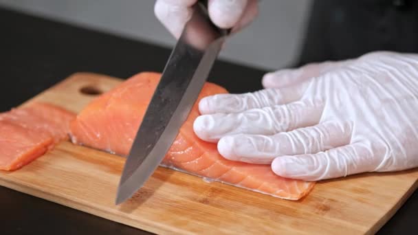 Un jeune chef sushi coupe un filet de saumon fumé avec un couteau sur une table. Mains dans les gants gros plan . — Video