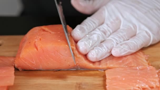 Un jeune chef sushi coupe un filet de saumon fumé avec un couteau sur une table. Mains dans les gants gros plan . — Video