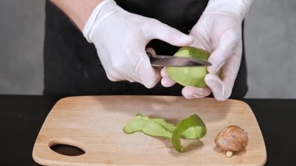 Young chef man in black clothes on a gray background peels, avocado with a knife. — Stock Video