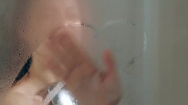 Duşta genç ve güzel bir kadın. Cam kabin. Sıcak duş, buhar ve duvarlarda damla. Bir parmak kalp çizer.