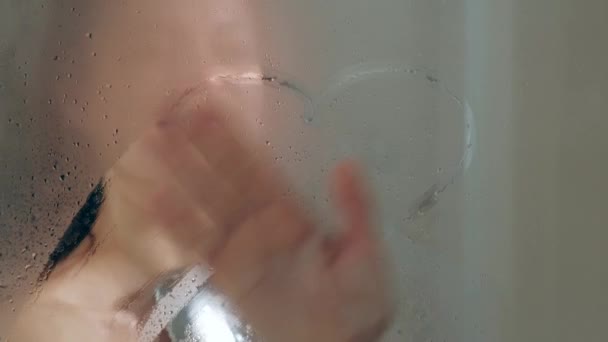 Joven mujer hermosa en la ducha. Cabina de cristal. Duchas calientes, vapor y gotas en las paredes. Dibuja un corazón de dedo . — Vídeos de Stock