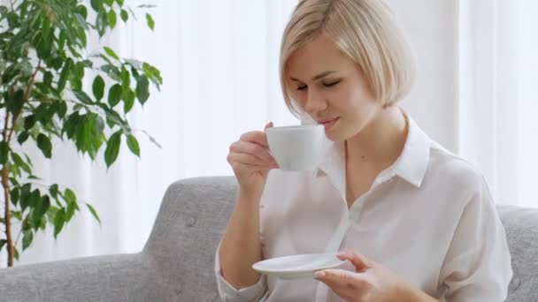 Uma jovem mulher loira bonita com cabelo curto com óculos bebe café de uma xícara branca em um apartamento leve . — Vídeo de Stock