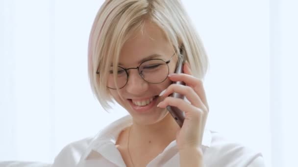 En ung vacker blond kvinna med kort hår glasögon pratar på en mobiltelefon när du sitter på en soffa i en ljus lägenhet. — Stockvideo