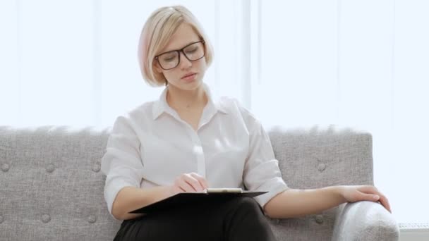 Eine junge schöne blonde Frau mit kurzen Haaren und Brille sitzt auf einem Sofa in einer hellen Wohnung. Arbeitsmüde, Hals- und Rückenschmerzen, Migräne. — Stockvideo