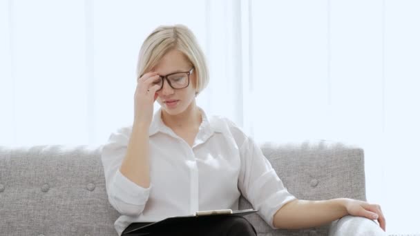 Uma mulher loira bonita nova com cabelo curto com óculos senta-se em um sofá em um apartamento leve. Cansado de trabalho, pescoço dolorido e costas, enxaqueca . — Vídeo de Stock