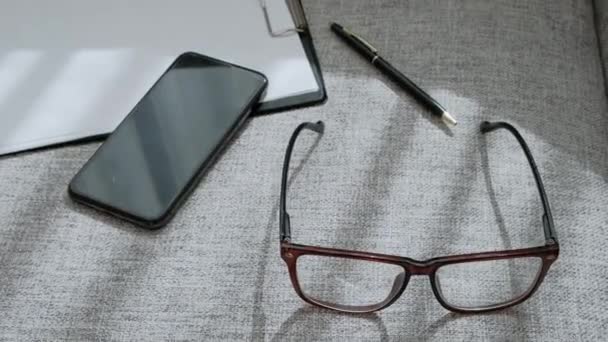 Óculos, telefone celular e notebook estão deitados no sofá. A mão feminina toma e põe óculos. Close-up, trabalho de escritório e negócios . — Vídeo de Stock