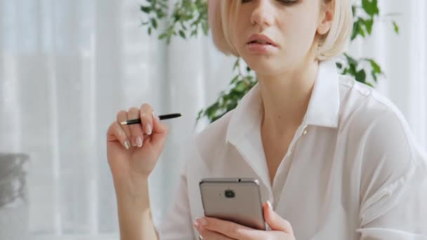 Uma jovem mulher loira bonita com cabelo curto vestindo óculos está falando em um telefone celular enquanto está sentado em um sofá em um apartamento brilhante . — Vídeo de Stock