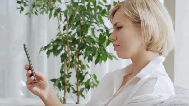 Une jeune belle femme blonde aux cheveux courts portant des lunettes parle sur un téléphone portable alors qu'elle est assise sur un canapé dans un appartement lumineux . — Video