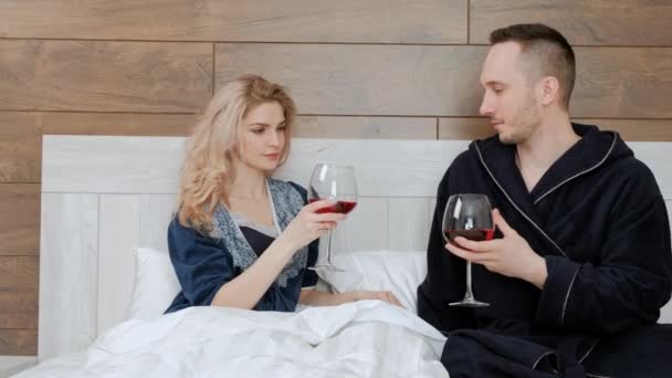 Jeune couple marié en peignoirs buvant du vin rouge couché sur un lit blanc dans la chambre de l'hôtel . — Video