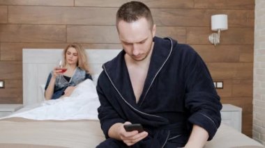 Bornozgenç bir evli çift otel odasında yatak odasında beyaz bir yatakta yatıyor. Bir adam akıllı telefonda konuşuyor, mesaj yazıyor. Bir kadın sıkılır ve şarap içer..