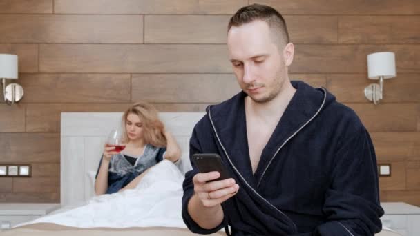 Um jovem casal de roupões de banho está deitado em uma cama branca no quarto do hotel. Um homem fala no smartphone, escreve mensagens. Uma mulher está entediada e bebe vinho . — Vídeo de Stock