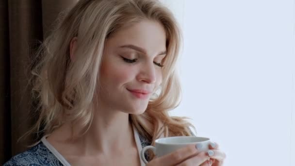 Una joven y hermosa rubia vestida con una túnica azul se encuentra junto a la ventana de una habitación y bebe té o café de una taza blanca. . — Vídeos de Stock