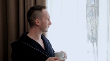 Bornozlu genç yakışıklı bir adam odanın penceresinde duruyor ve beyaz bir fincandan çay ya da kahve içiyor..