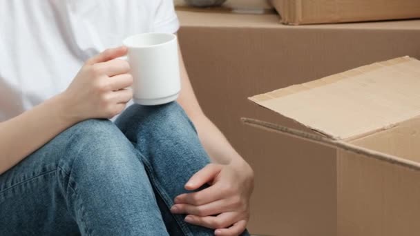 Jonge mooie vrouw in jeans en een wit T-shirt is het drinken van koffie terwijl zittend op de vloer tegen een achtergrond van kartonnen dozen. — Stockvideo