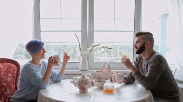 Jong mooi paar in warme truien in een landhuis zitten aan de witte tafel tegen de achtergrond van het raam. De familie drinkt thee uit witte kopjes. — Stockvideo