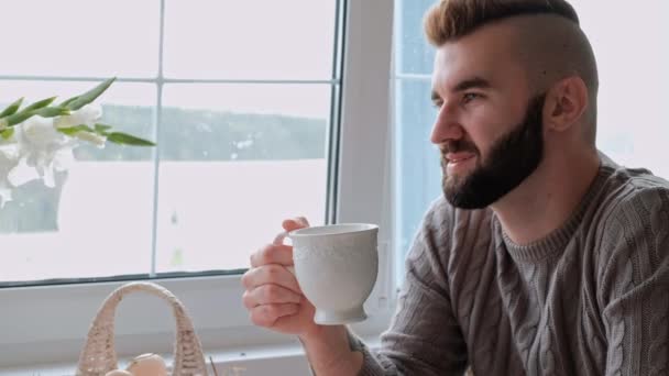 Jeune beau couple en pull chaud dans une maison de campagne sont assis à la table blanche sur le fond de la fenêtre. La famille boit du thé dans des tasses blanches . — Video