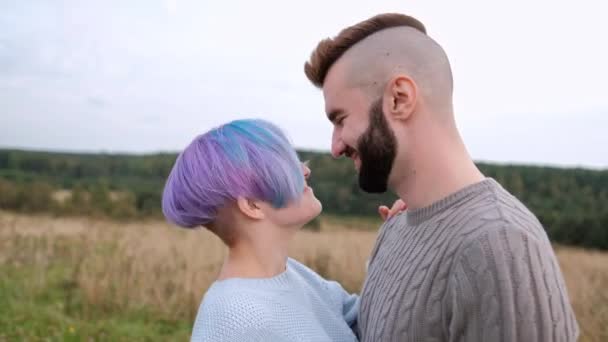 Joven pareja hermosa en suéteres y sombreros contra el fondo de la naturaleza, campos con hierba verde y bosque. Cabello corto y teñido . — Vídeo de stock