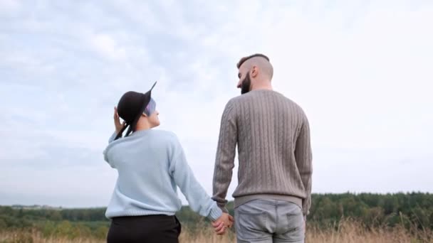 Joven pareja hermosa en suéteres y sombreros contra el fondo de la naturaleza, campos con hierba verde y bosque. Cabello corto y teñido . — Vídeo de stock