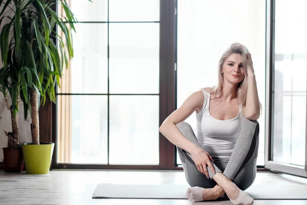 Joven hermosa atlética rubia en ropa deportiva blanca haciendo yoga y estirándose sobre una alfombra gris en el fondo de una gran ventana de edificio de varios pisos . — Foto de Stock