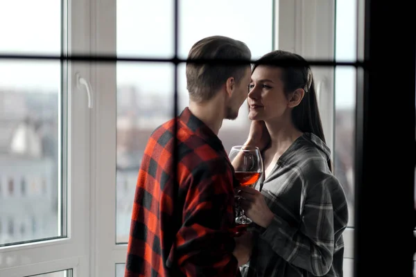 Jovem casal bonito em camisas xadrez na varanda de um edifício de vários andares. Beber e provar vinho, rir e abraçar . — Fotografia de Stock