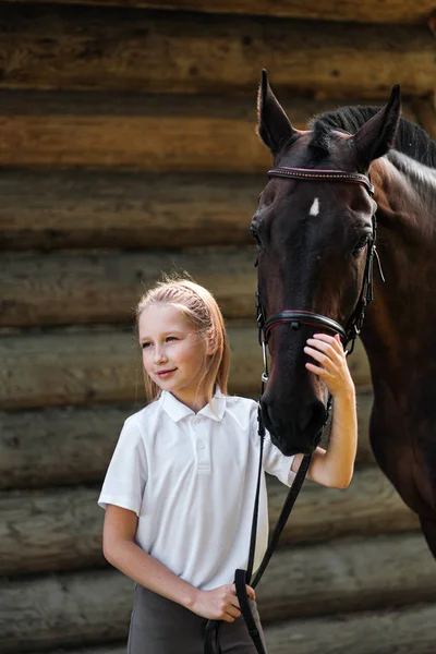 Una jinete adolescente se para junto a un caballo marrón y la abraza. Sobre el fondo de un establo de madera . — Foto de Stock