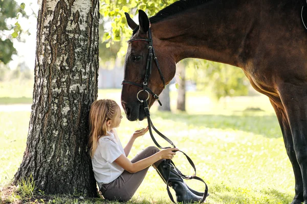 Chica jinete adolescente se sienta en un claro verde bajo un árbol. Alimenta a un caballo con una manzana y lo acaricia . — Foto de Stock