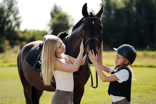 Madre e hija jinetes y jinetes en un campo verde abrazan a un caballo marrón . — Foto de Stock