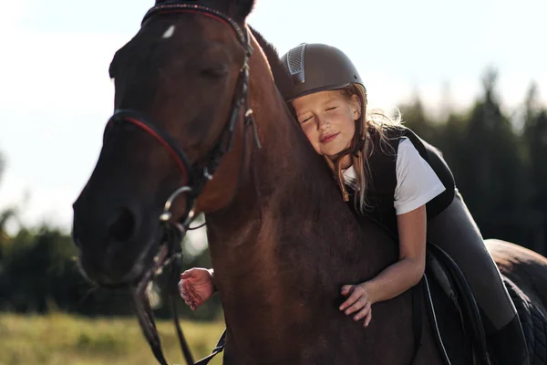 Menina jockey adolescente senta-se em um cavalo marrom, abraços e acaricia-la . — Fotografia de Stock