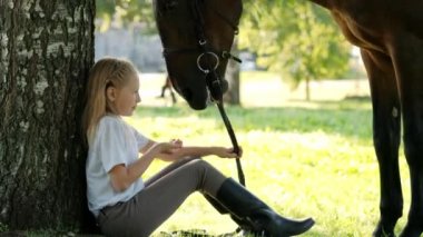 Genç kız jokey bir ağacın altındaki yeşil açıklıkta oturuyor. Bir ata elma yedirir ve onu okşar..