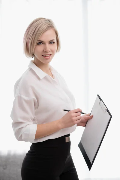 Menina loira bonita nova com cabelo curto em uma camisa branca está de pé no escritório e segurando um caderno e caneta . — Fotografia de Stock