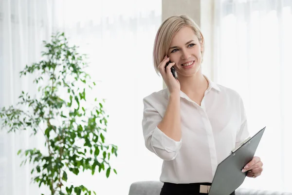 Jong mooi blond meisje met kort haar in een wit overhemd staat fel in het kantoor tegen het raam. Bevat een notebook en smartphone. Praten over de telefoon. — Stockfoto