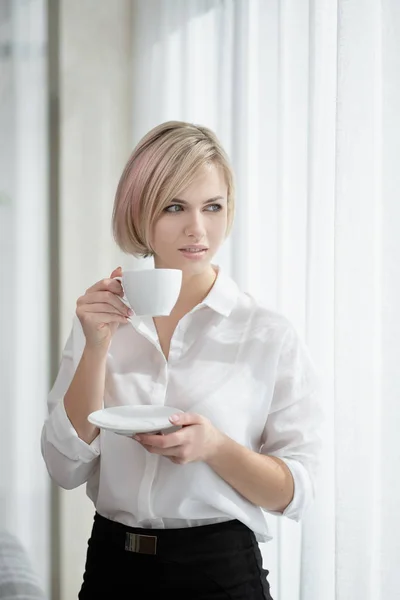 Νέα όμορφη ξανθιά κορίτσι με κοντά μαλλιά σε ένα λευκό πουκάμισο και γυαλιά κάθεται στον καναπέ στο φως στο γραφείο πάνω από το παράθυρο. Κρατά ένα λευκό φλιτζάνι και πιατάκι. Πίνοντας καφέ ή τσάι σε ένα — Φωτογραφία Αρχείου