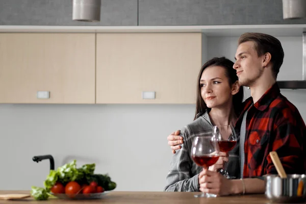 Beautiful young couple in plaid shirts in the kitchen preparing food and drinking red wine from glasses. — Stock Photo, Image