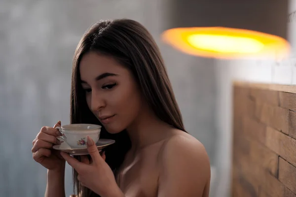 Piękna brunetka dziewczyna z brązowymi oczami z gołymi ramionami pije herbatę lub kawę z małej filiżanki porcelany w sypialni na łóżku. — Zdjęcie stockowe